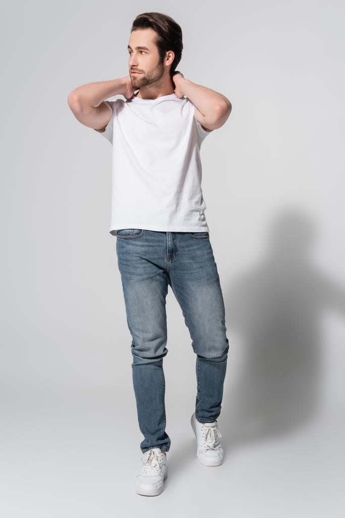 Ganzkörperansicht eines jungen Mannes in Jeans und weißem T-Shirt, der mit den Händen hinter dem Hals auf grau posiert - Foto, Bild