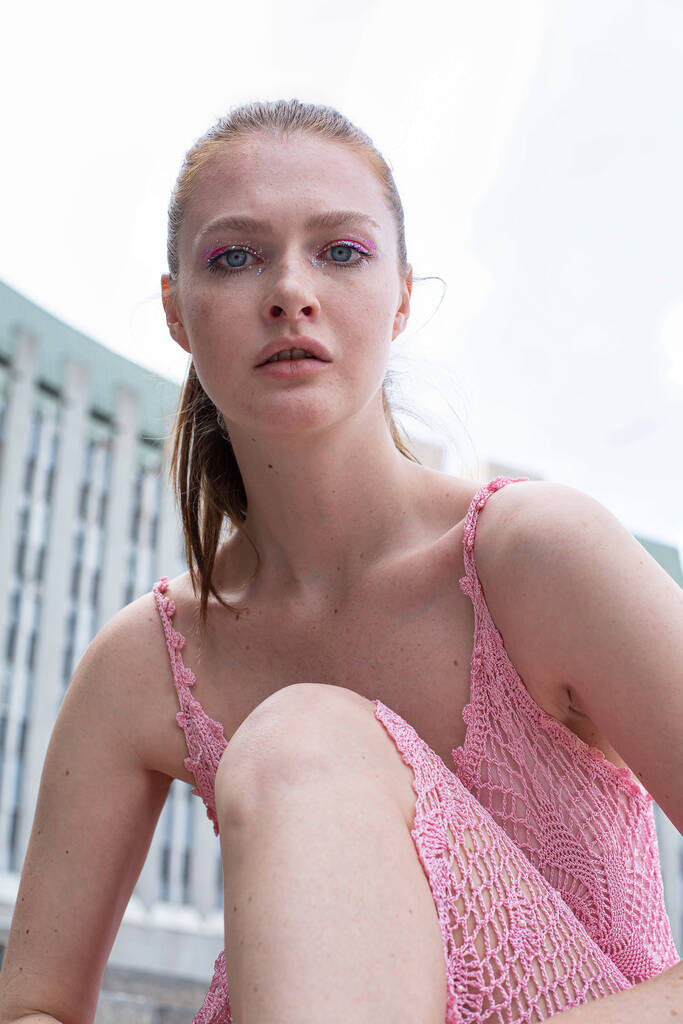 White blond young woman with a ponytail in pink summer dress with blue eyes with building behind - Photo, Image