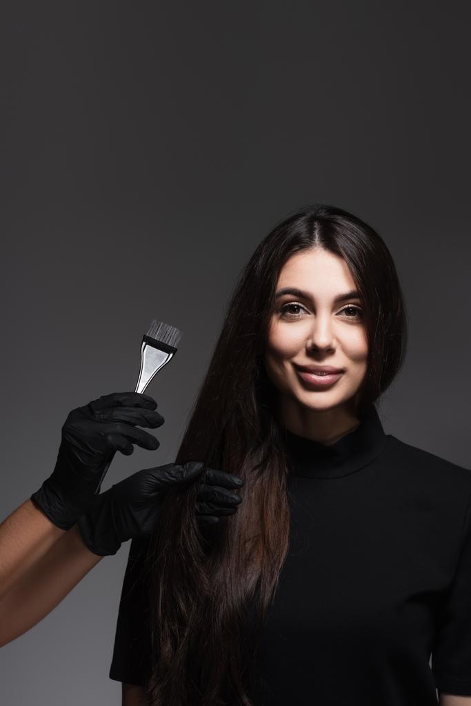 kadeřnice drží štětec poblíž veselá žena s hnědými vlasy izolované na tmavě šedé - Fotografie, Obrázek