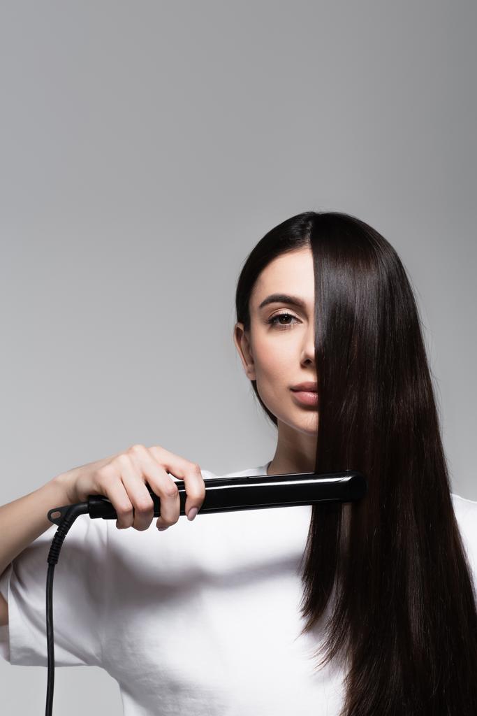 mladá brunetka žena pomocí rovnač vlasů izolované na šedé - Fotografie, Obrázek