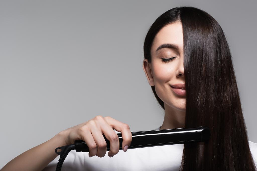 довольная брюнетка женщина с помощью выпрямителя волос изолированы на сером - Фото, изображение