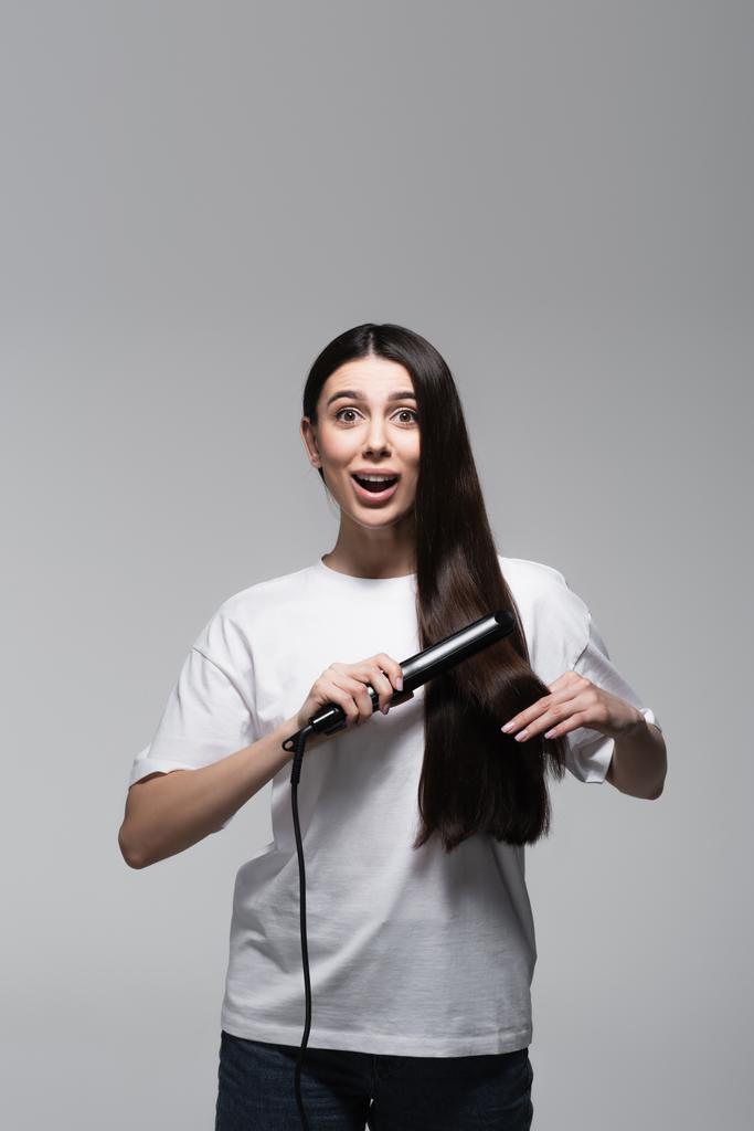 молодая изумленная женщина, использующая выпрямитель для волос, изолированный на сером - Фото, изображение
