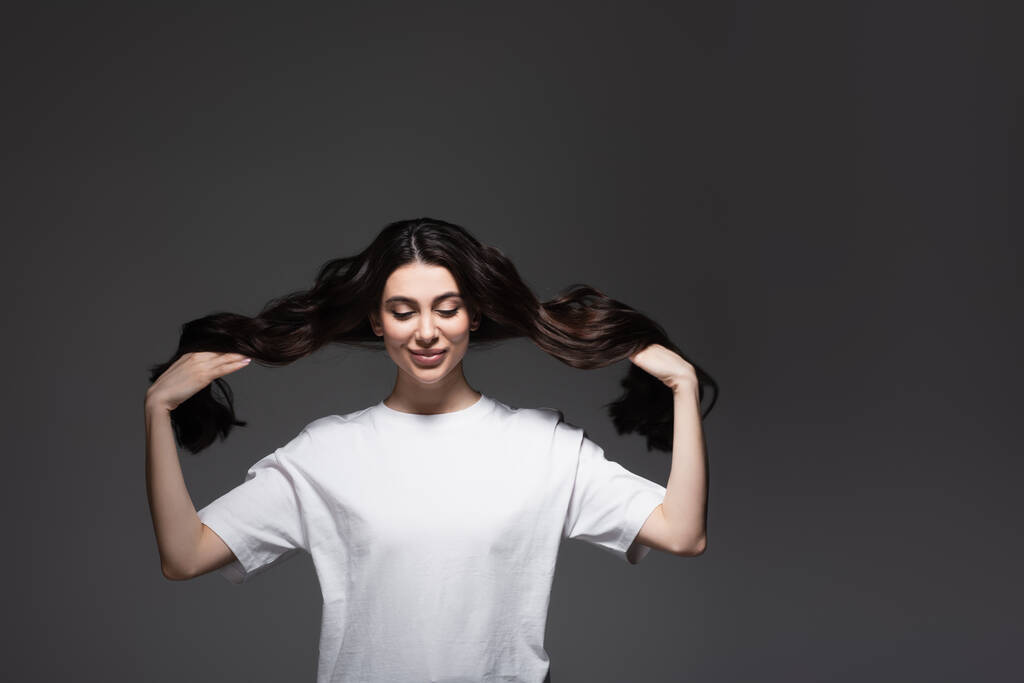 nuori iloinen nainen säätää pitkiä aaltoilevia hiuksia tummanharmaalla  - Valokuva, kuva