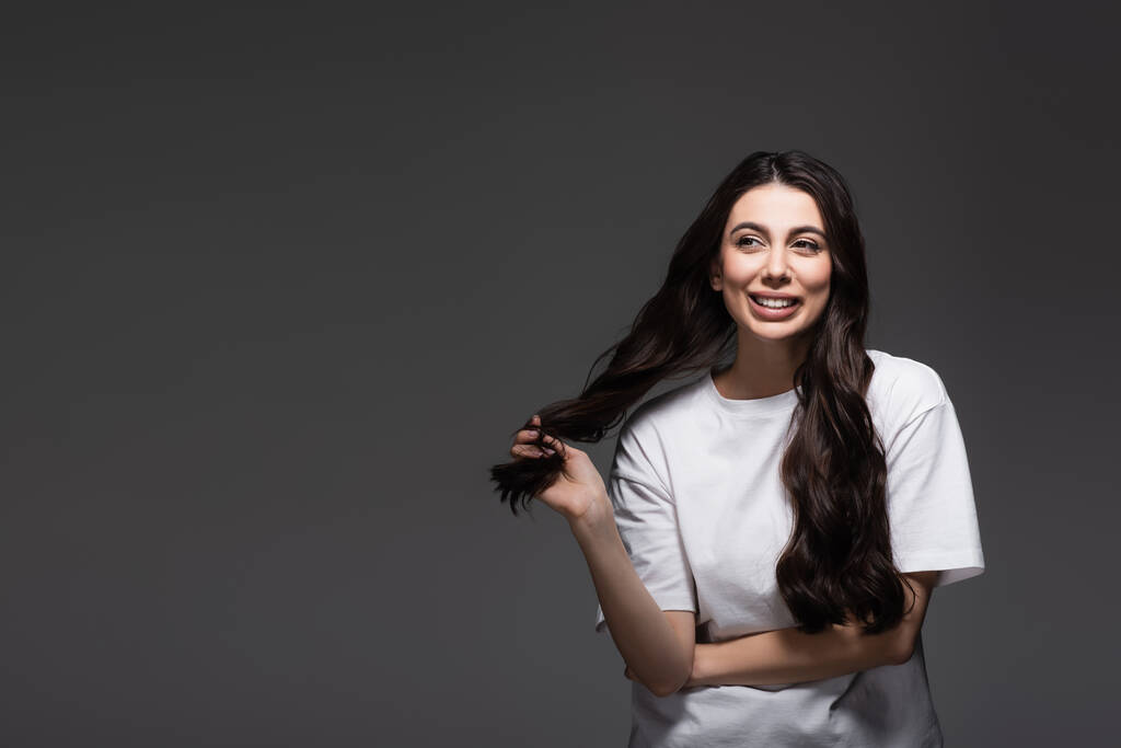 молодая счастливая женщина регулирует волнистые волосы на темно-сером  - Фото, изображение