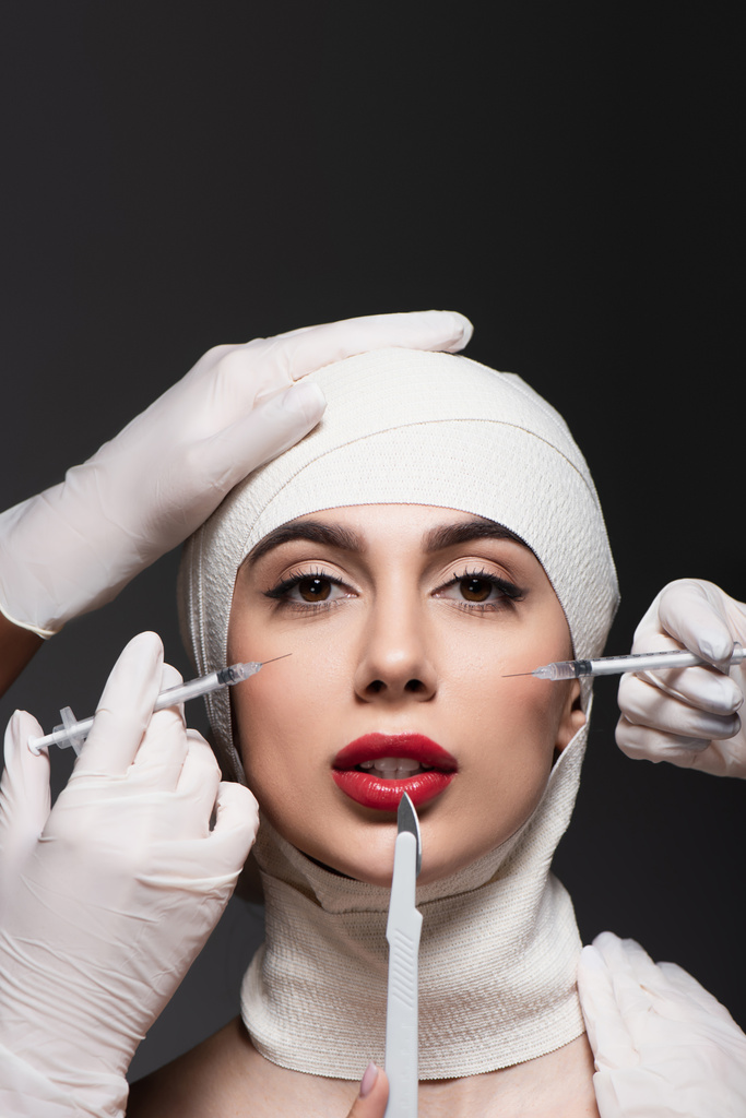 chirurgiens plasticiens en gants de latex tenant un couteau chirurgical et des seringues près de la femme en bandage élastique sur la tête isolé sur gris foncé  - Photo, image