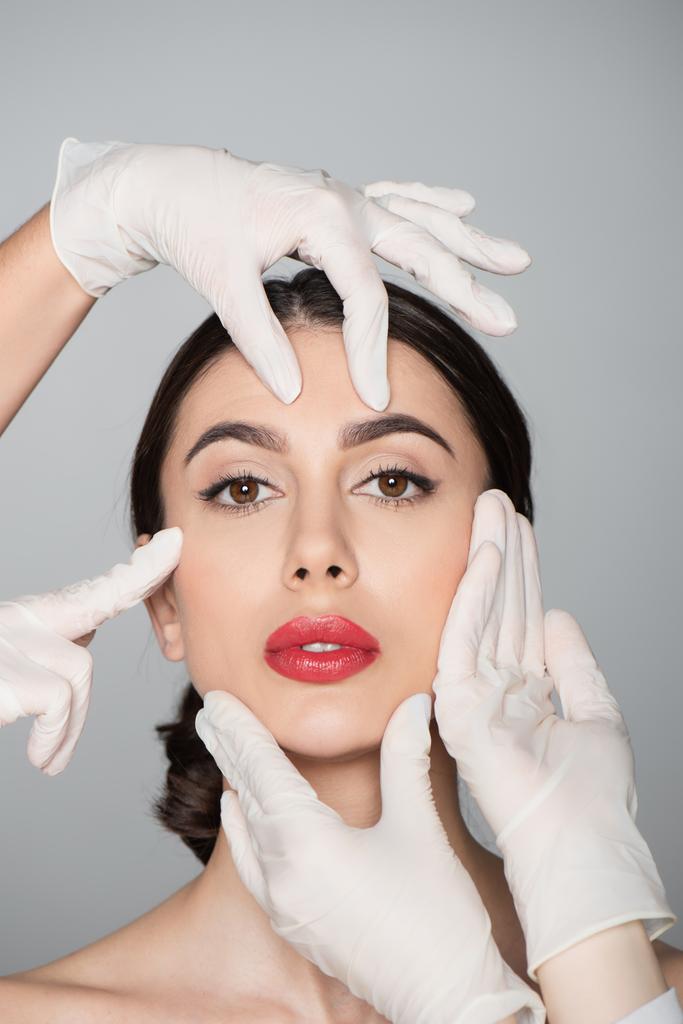 Plastische Chirurgen in Latex-Handschuhen berühren Gesicht der jungen Frau isoliert auf grau - Foto, Bild