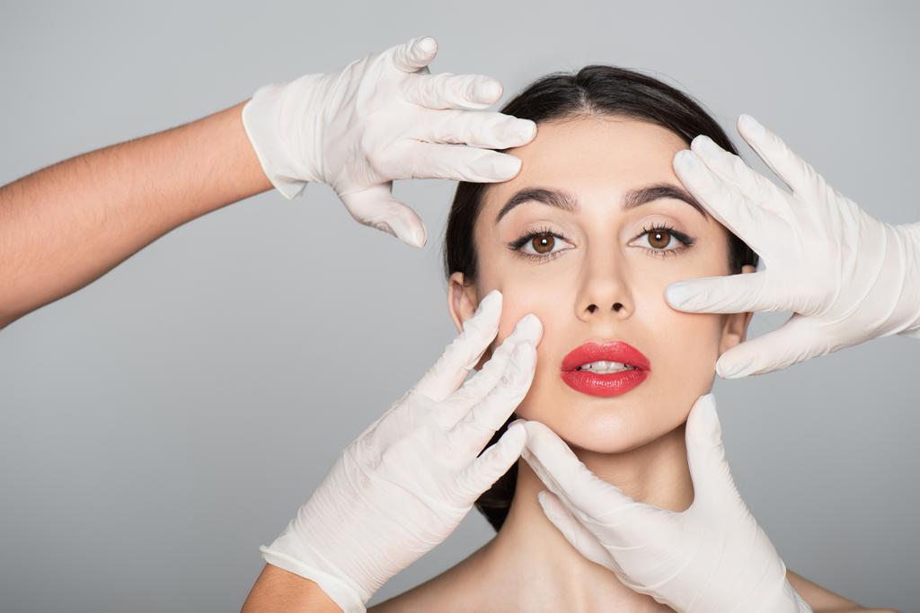 Пластические хирурги в латексных перчатках трогают лицо брюнетки женщины, изолированной на серой - Фото, изображение