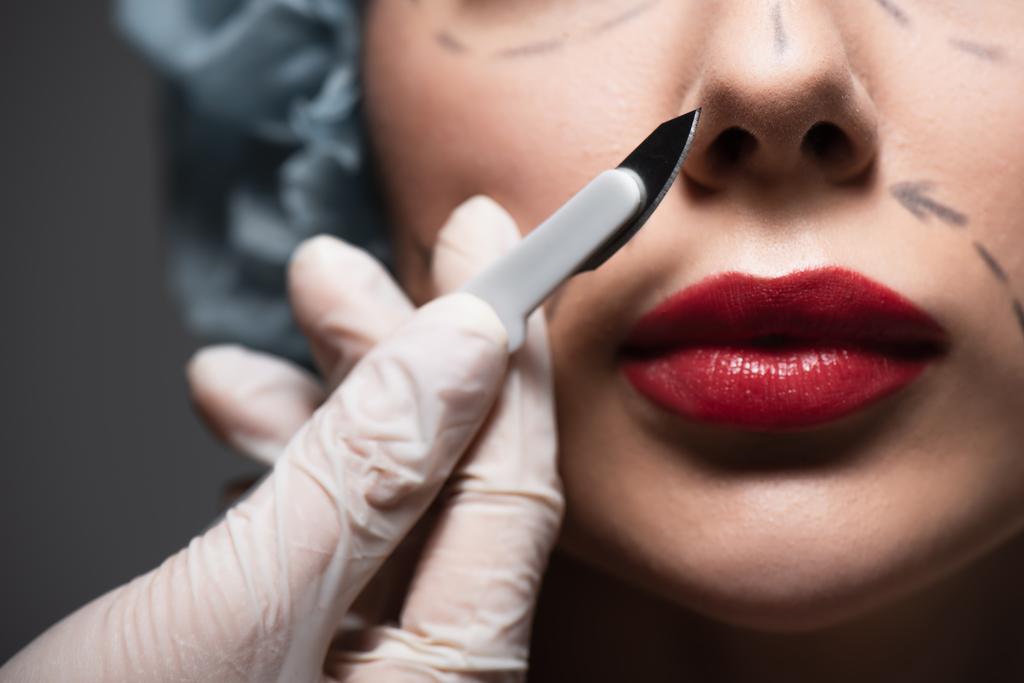 close up of plastic surgeon in latex glove holding scalpel near woman with marked lines on face isolated on grey - Photo, Image