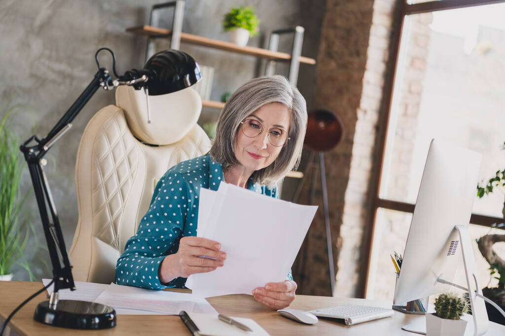Фото спокойной серьезной ориентированной старой деловой женщины выглядят документы офисная работа сидеть стол в помещении на рабочем месте - Фото, изображение