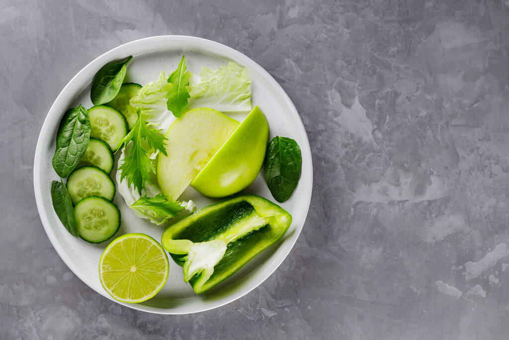 Gri arka planda dilimlenmiş yeşil meyve ve sebzeler. Beyaz tabakta taze yeşil diyet yemeği. Sağlıklı vejetaryen gıda konsepti. Uzayı kopyala Üst görünüm - Fotoğraf, Görsel