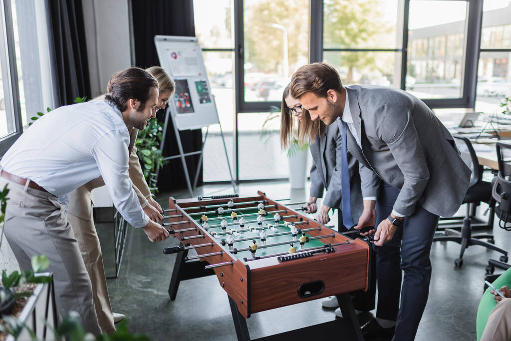 mladí a šťastní podnikatelé ve formálním oblečení hrát stolní fotbal v kanceláři - Fotografie, Obrázek