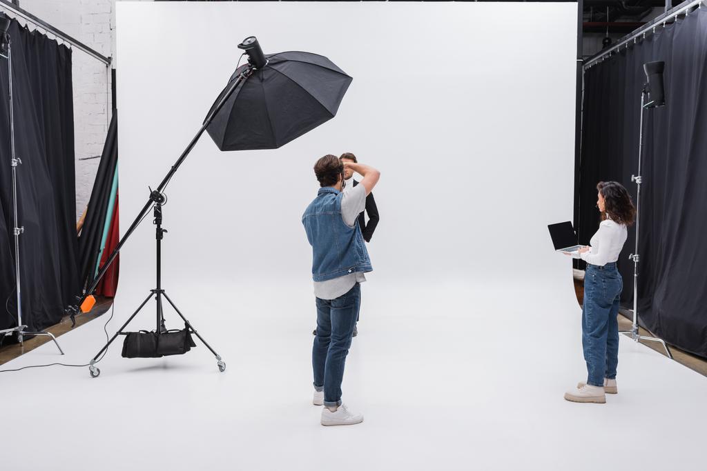 directeur artistique tenant ordinateur portable avec écran vierge près du photographe et du modèle travaillant dans un studio photo - Photo, image