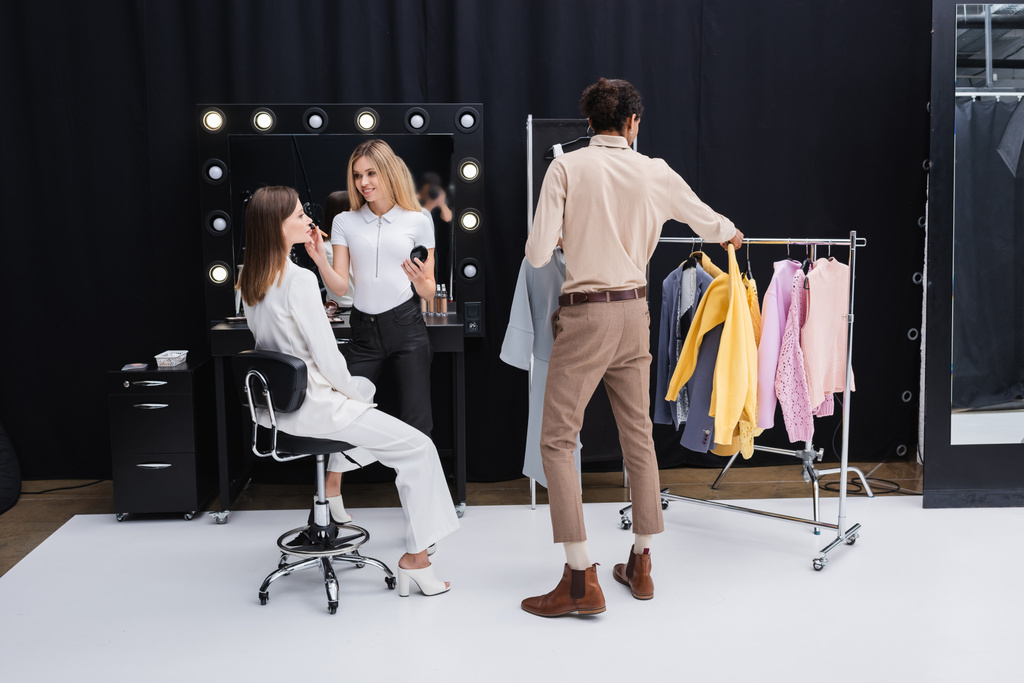 african american stylist choosing clothes while makeup artist doing visage to model in dressing room - Photo, Image
