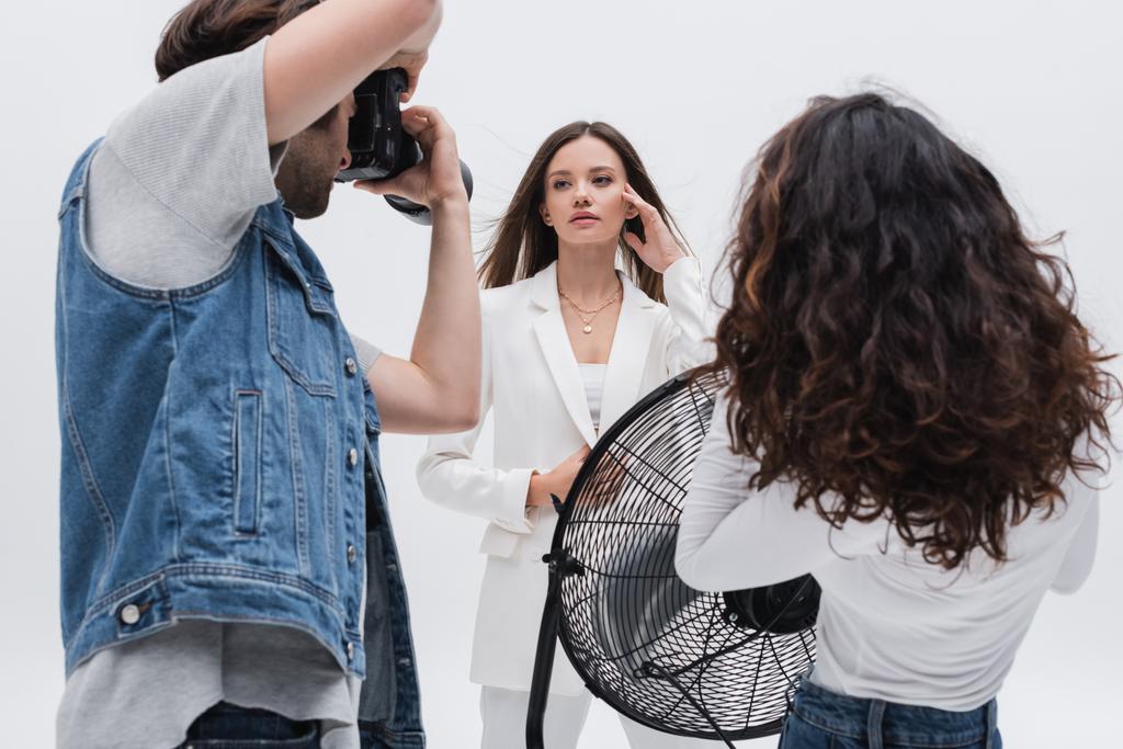 ブルネット女性でホワイトスーツのポーズ近くに写真家とアシスタントとともに電動ファン分離オンホワイト - 写真・画像