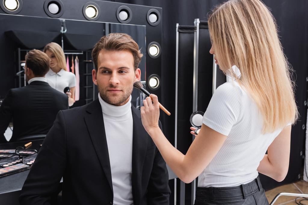 rubia visage artista aplicación de maquillaje en el hombre cerca del espejo en estudio de fotos - Foto, imagen