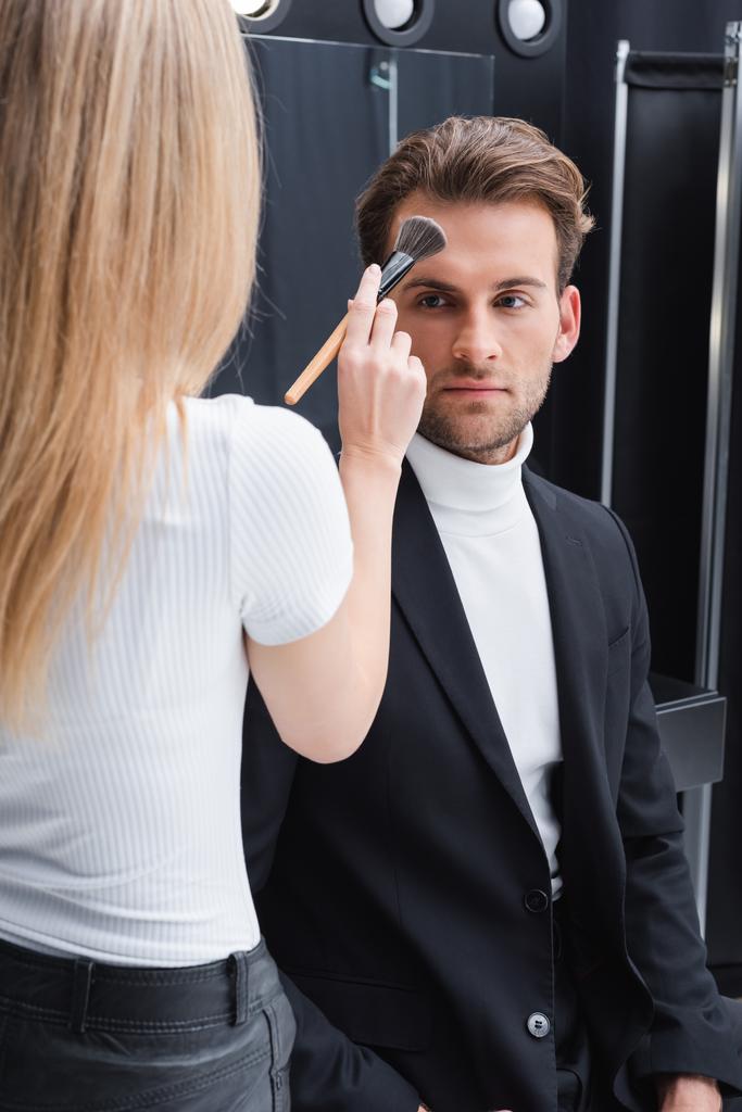 widok z tyłu blondynka twarz artysta stosowania makijaż na mężczyzna w studio fotograficznym - Zdjęcie, obraz