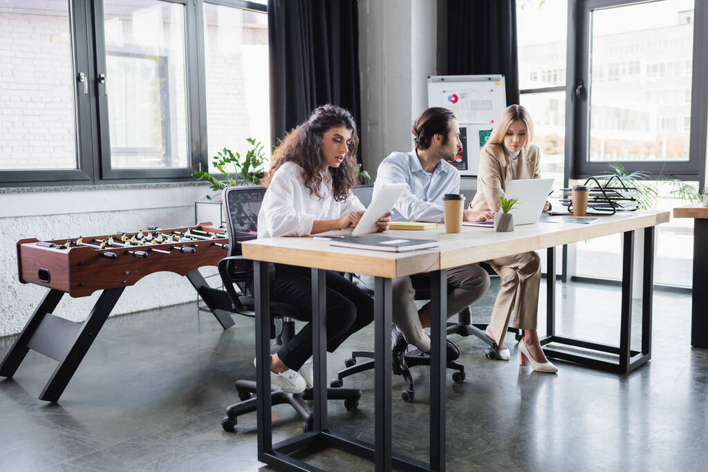молодые бизнесмены сидят за столом с устройствами, работая вместе в офисе - Фото, изображение