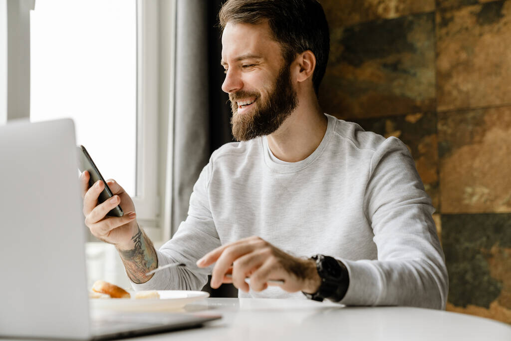 Homme barbu souriant et utilisant un téléphone portable tout en travaillant avec un ordinateur portable à l'intérieur - Photo, image