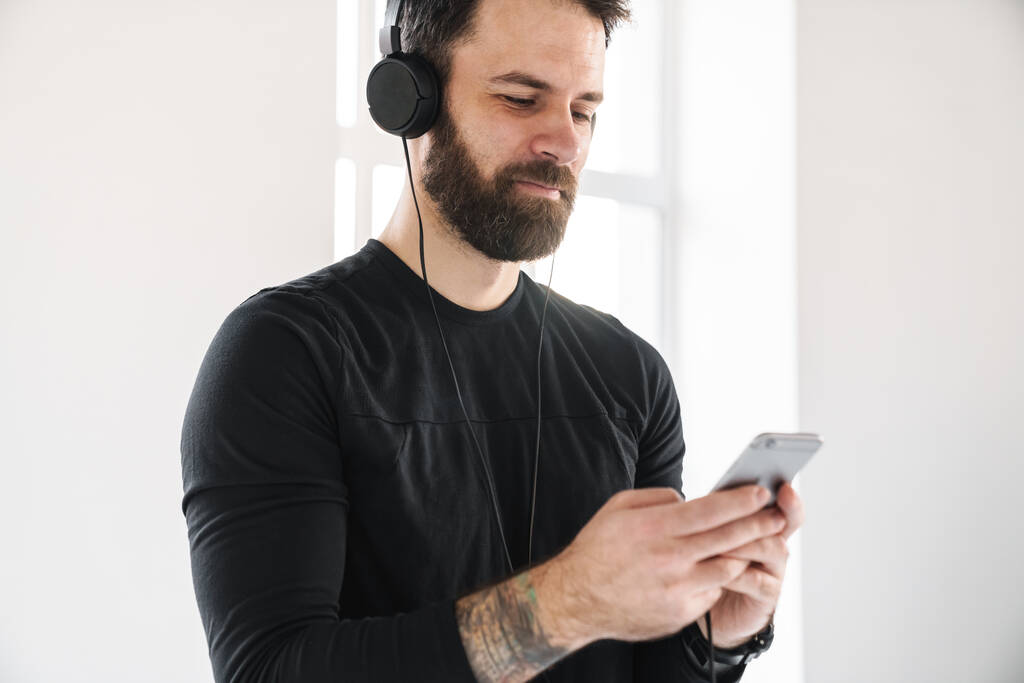 Jonge brunette bebaarde blanke man in sportkleding binnen staan luisteren naar muziek in koptelefoon met mobiele telefoon - Foto, afbeelding