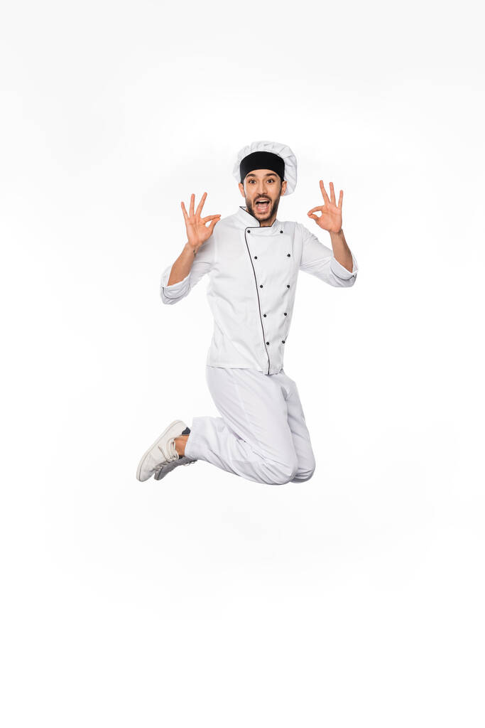 схвильований шеф-кухар у капелюсі та уніформі, що стрибає та показує добре на білому
  - Фото, зображення