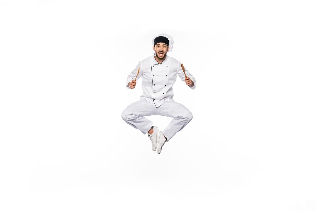 allegro chef in cappello e uniforme saltando e tenendo in mano spatola in legno e cucchiaio isolato su bianco  - Foto, immagini