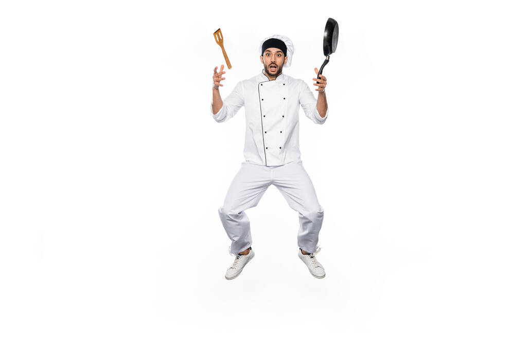 Chef sorprendido en sombrero y uniforme levitando cerca de la sartén voladora con espátula de madera aislada en blanco  - Foto, Imagen