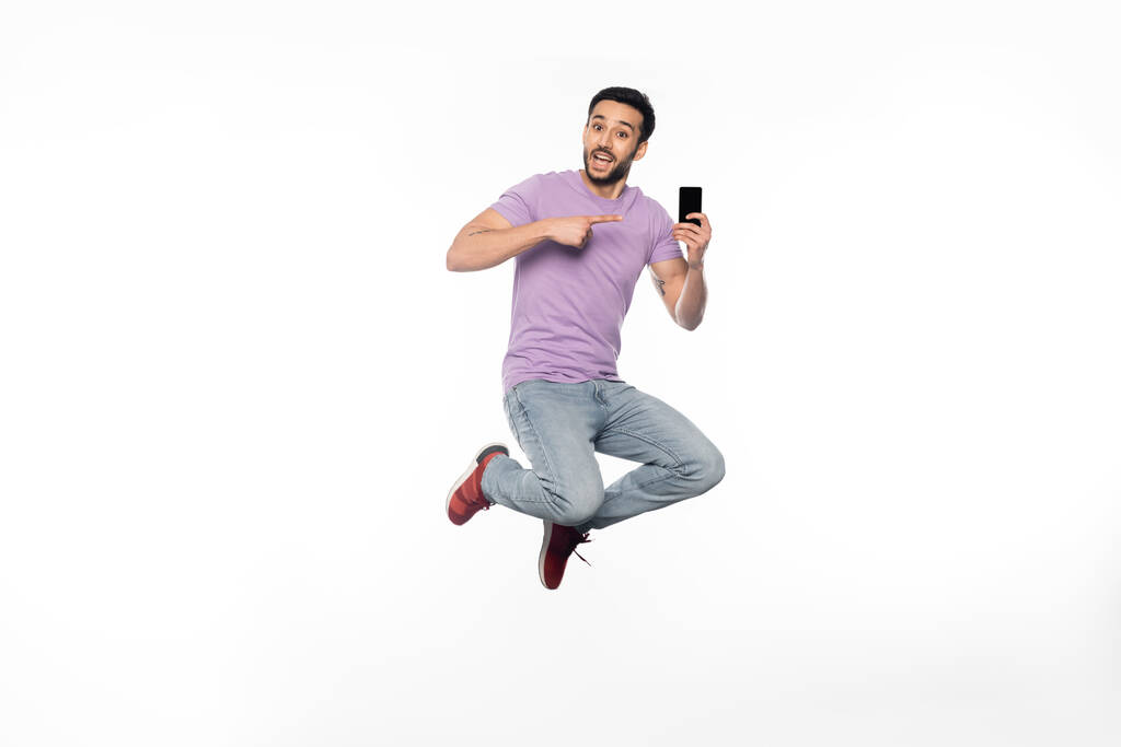 κατάπληκτος άνθρωπος με τζιν και μωβ t-shirt αιωρείται ενώ δείχνει στο smartphone σε λευκό  - Φωτογραφία, εικόνα