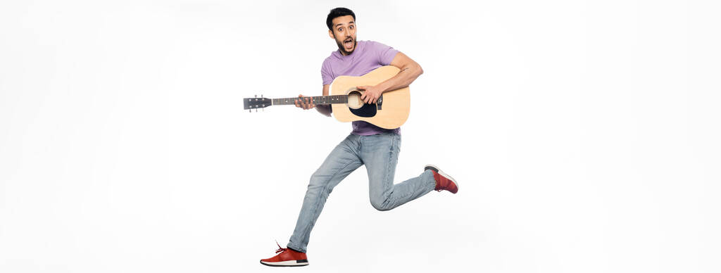 Verblüffter Mann in Jeans und lila T-Shirt schwebt während er Akustikgitarre auf weißem Banner spielt - Foto, Bild