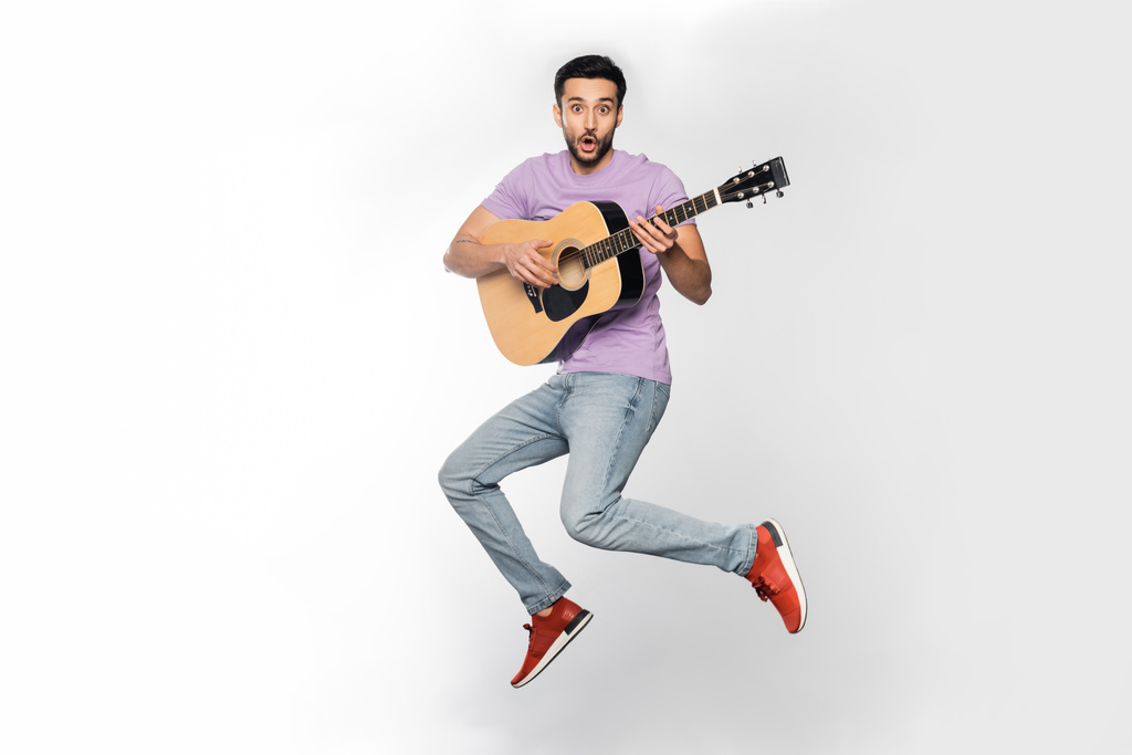 Schockierter Mann in Jeans und lila T-Shirt schwebt, während er Akustikgitarre auf weißem Hintergrund spielt  - Foto, Bild