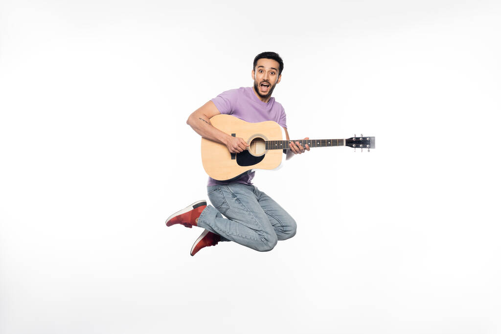 hombre positivo en jeans y camiseta púrpura levitando mientras toca la guitarra acústica en blanco  - Foto, imagen