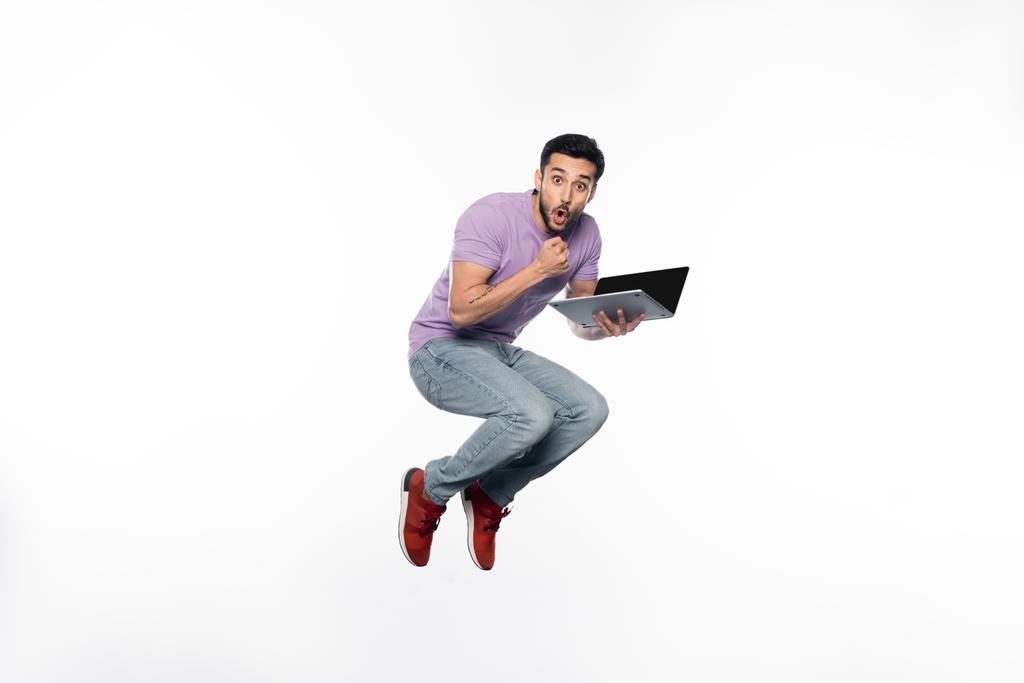 zszokowany mężczyzna w dżinsach i fioletowy t-shirt lewitujący trzymając laptopa na białym  - Zdjęcie, obraz