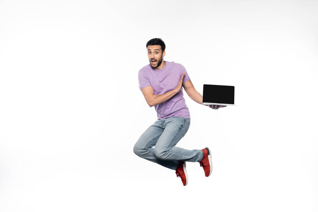 Erstaunter Mann in Jeans und lila T-Shirt schwebt, während er Laptop auf weißem Hintergrund hält  - Foto, Bild