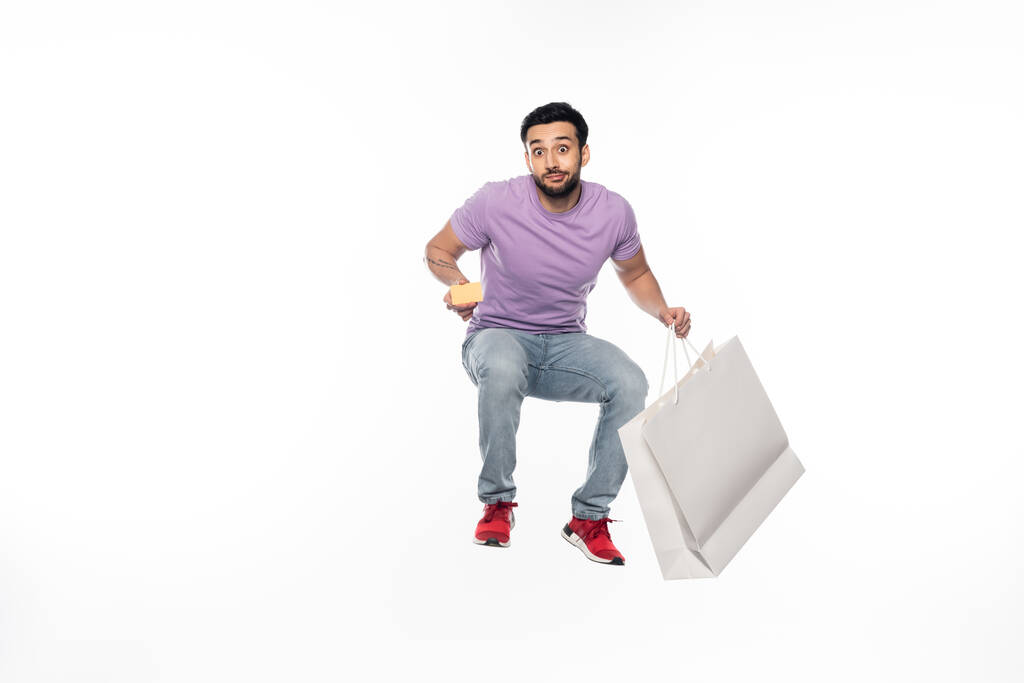 Mann in Jeans und lila T-Shirt schwebt, während er Kreditkarte und Einkaufstasche auf weiß hält - Foto, Bild