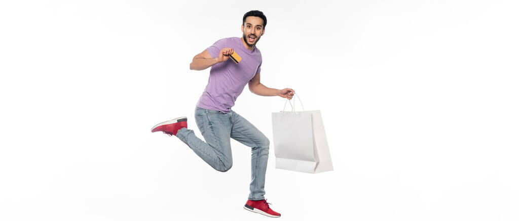 homme excité en jeans et t-shirt violet lévitant tout en tenant la carte de crédit et le sac à provisions sur blanc, bannière - Photo, image
