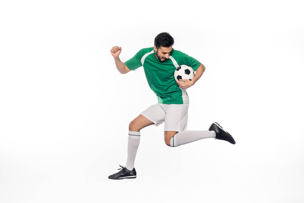 νεαρός ποδοσφαιριστής με στολή άλμα με μπάλα ποδοσφαίρου σε λευκό  - Φωτογραφία, εικόνα