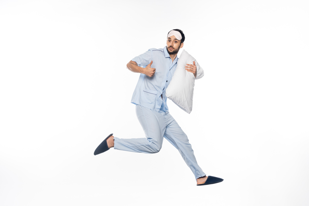 bearded man in pajamas and sleeping mask levitating with pillow while showing thumb up on white  - Photo, Image