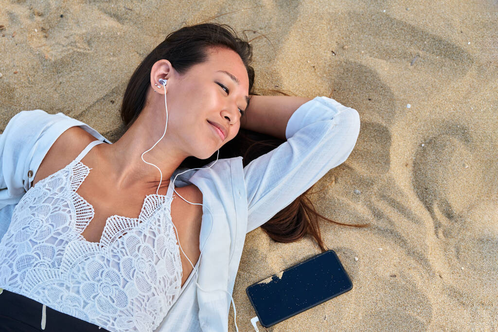 Расслабляющая молодая женщина в наушниках со смартфоном на песке, вид сверху - Фото, изображение
