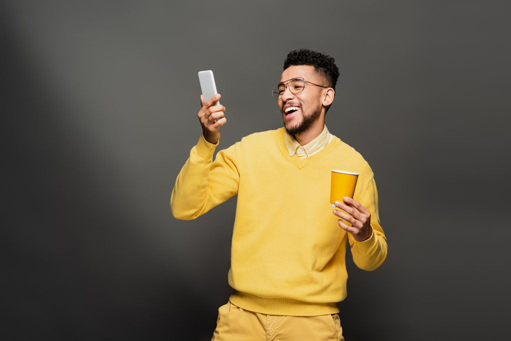 счастливый африканский американец в очках и желтом свитере, держащий бумажную чашку и смотрящий на смартфон на темно-сером - Фото, изображение