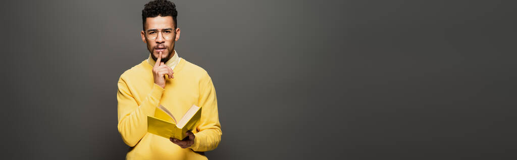 hombre afroamericano reflexivo en gafas y traje amarillo sosteniendo libro en gris oscuro, bandera - Foto, imagen