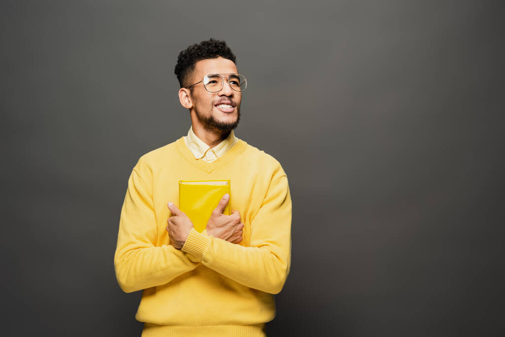 smiling african american man in glasses and yellow outfit holding book on dark grey - Photo, Image