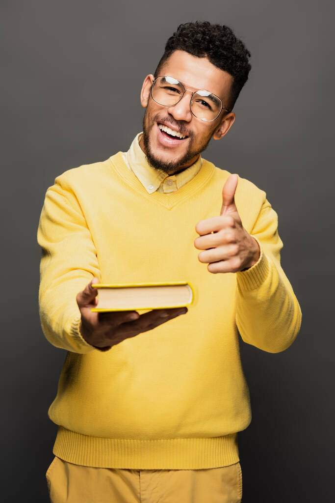 happy african american man in glasses and yellow sweater showing thumb up and holding book isolated on grey - Photo, Image