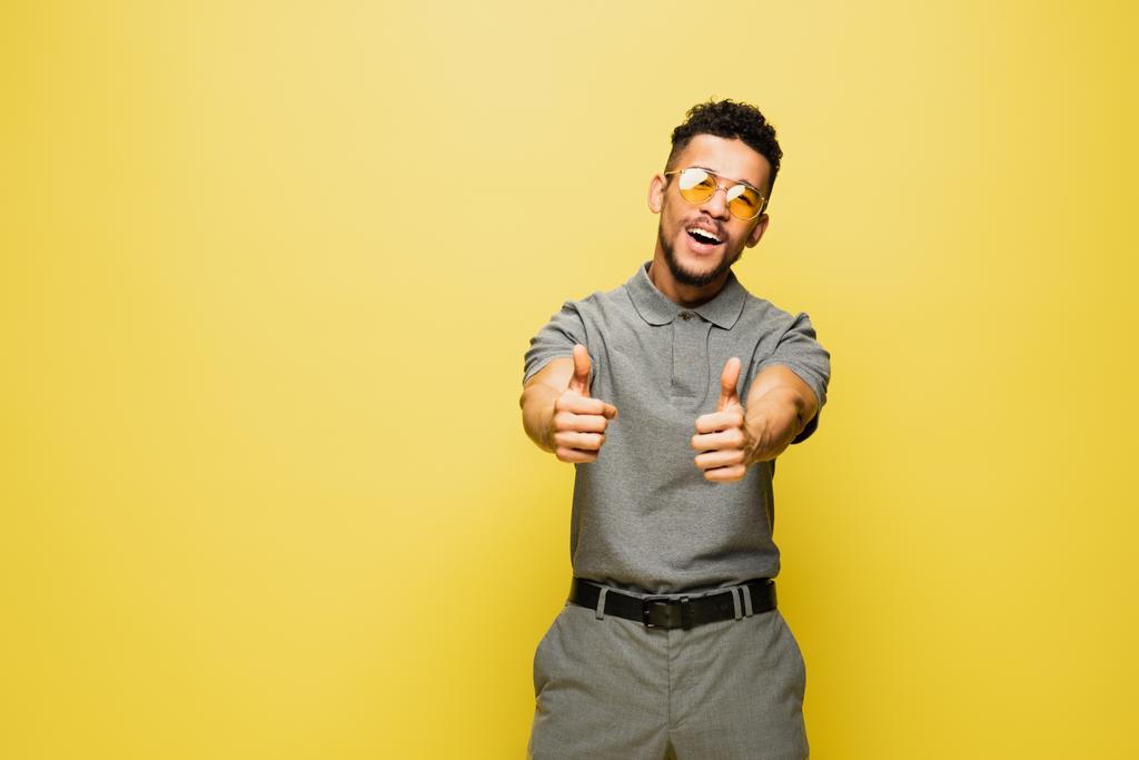 radosny Afrykański Amerykanin w okularach przeciwsłonecznych i szarej koszuli tenisowej z kciukami w górze na żółtym  - Zdjęcie, obraz