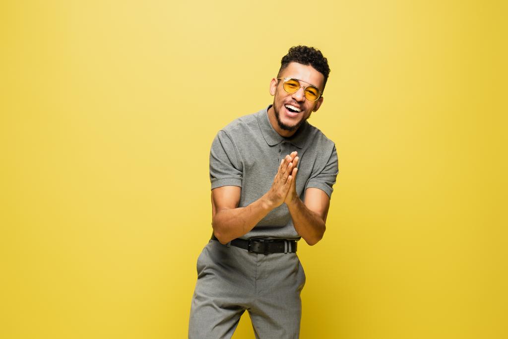 pozitív afro-amerikai férfi napszemüvegben és szürke tenisz ing imádkozó kézzel elszigetelt sárga  - Fotó, kép