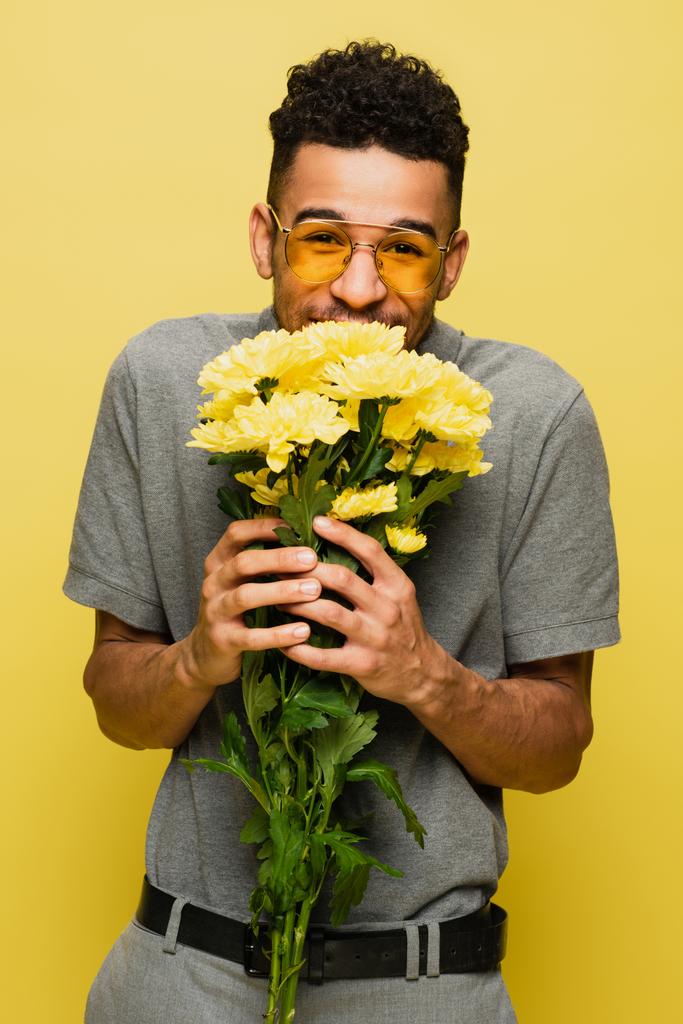 šťastný africký Američan v slunečních brýlích a šedé tenisové košili drží kytice květin izolovaných na žluté  - Fotografie, Obrázek