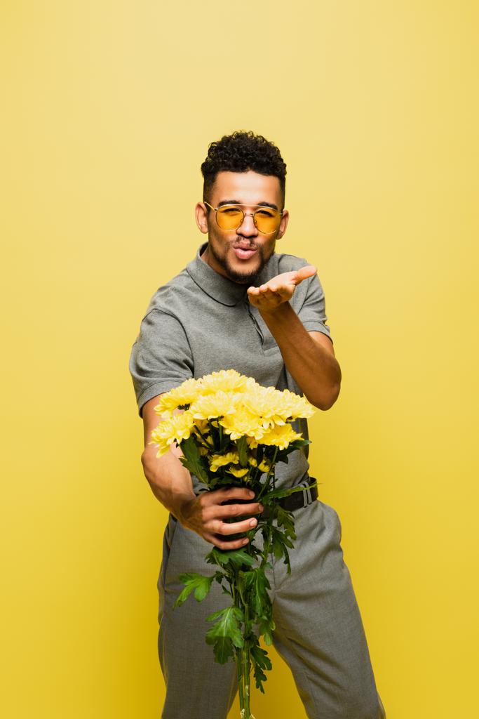 stylische afrikanisch-amerikanische Mann mit Sonnenbrille hält Blumenstrauß und sendet Luftkuss isoliert auf gelb  - Foto, Bild
