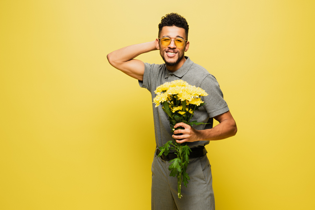 šťastný africký Američan v slunečních brýlích drží kytice květin na žluté  - Fotografie, Obrázek