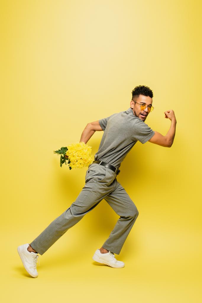 longitud completa de hombre afroamericano en gafas de sol con ramo de flores y corriendo en amarillo  - Foto, Imagen
