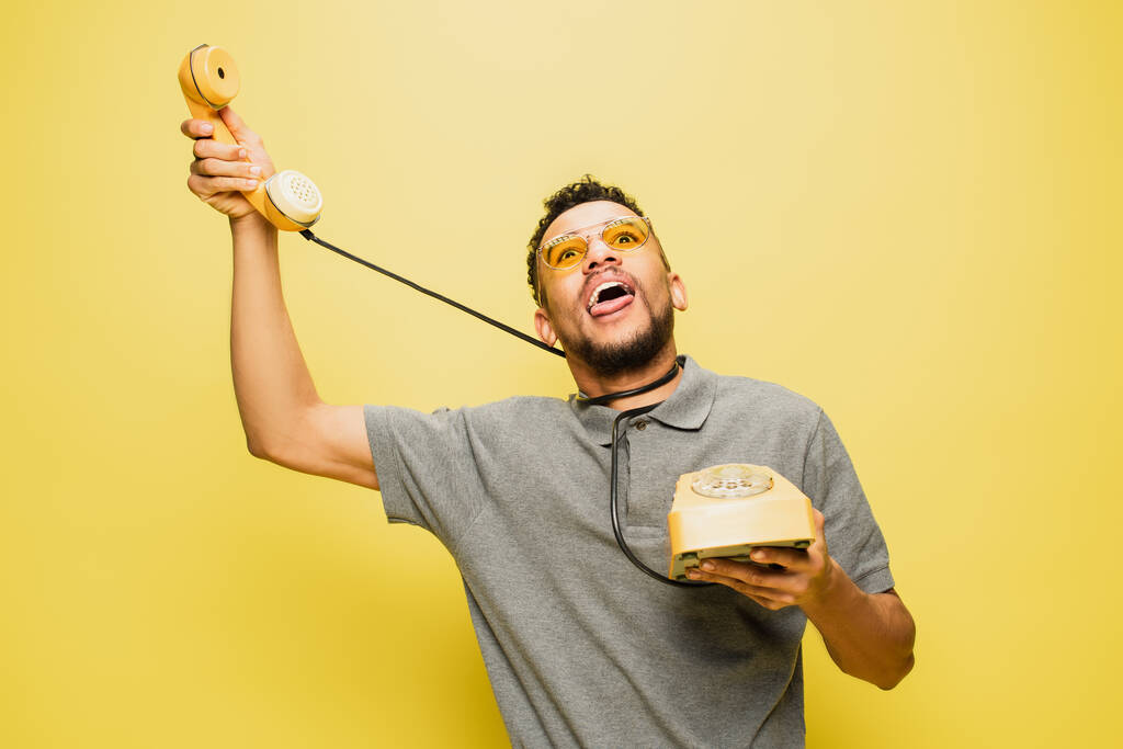 młody Afrykanin amerykański człowiek w okularach przeciwsłonecznych wystający język podczas duszenia się kablem telefonicznym na żółty  - Zdjęcie, obraz