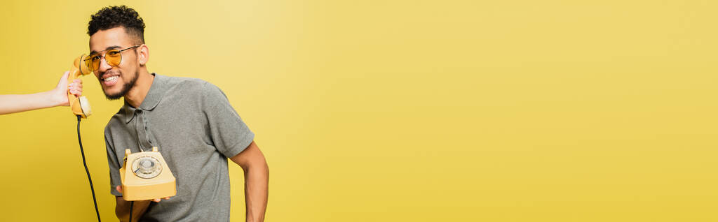 黄色のバナーにサングラスとグレーのテニスシャツを着た陽気なアフリカ系アメリカ人男性の近くに携帯電話を持っている女性 - 写真・画像