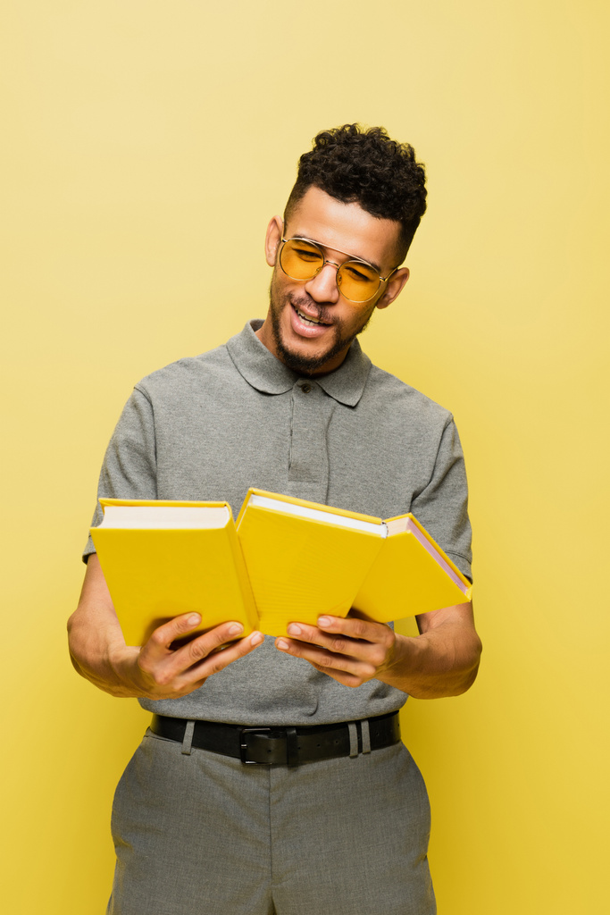 Χαρούμενος Αφροαμερικάνος με γυαλιά ηλίου και γκρι πουκάμισο τένις που κρατάει βιβλία απομονωμένα στο κίτρινο.  - Φωτογραφία, εικόνα